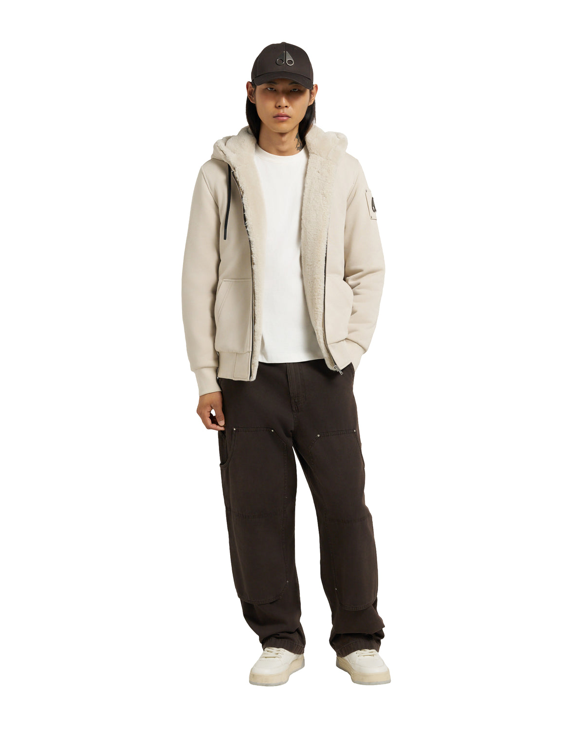 A person wearing a hooded zip-up jacket, a white t-shirt, dark pants, white sneakers, and a baseball cap.