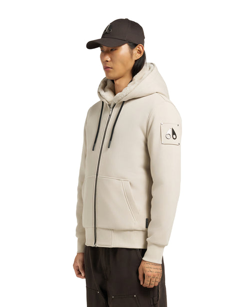 A person wearing a cream-colored hoodie with a full zipper, a hood, and a logo patch on the left sleeve.