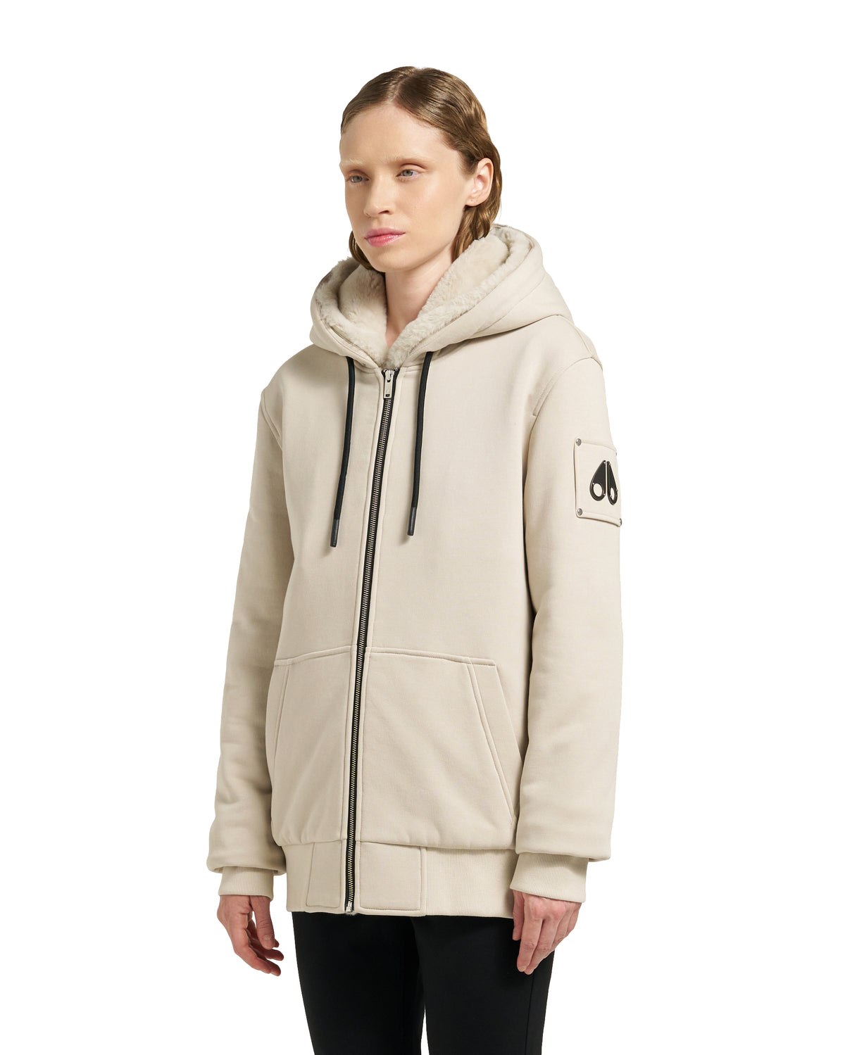 A woman models a beige hoodie with a black zipper and drawstring, featuring the moose knuckle logo on the left sleeve.
