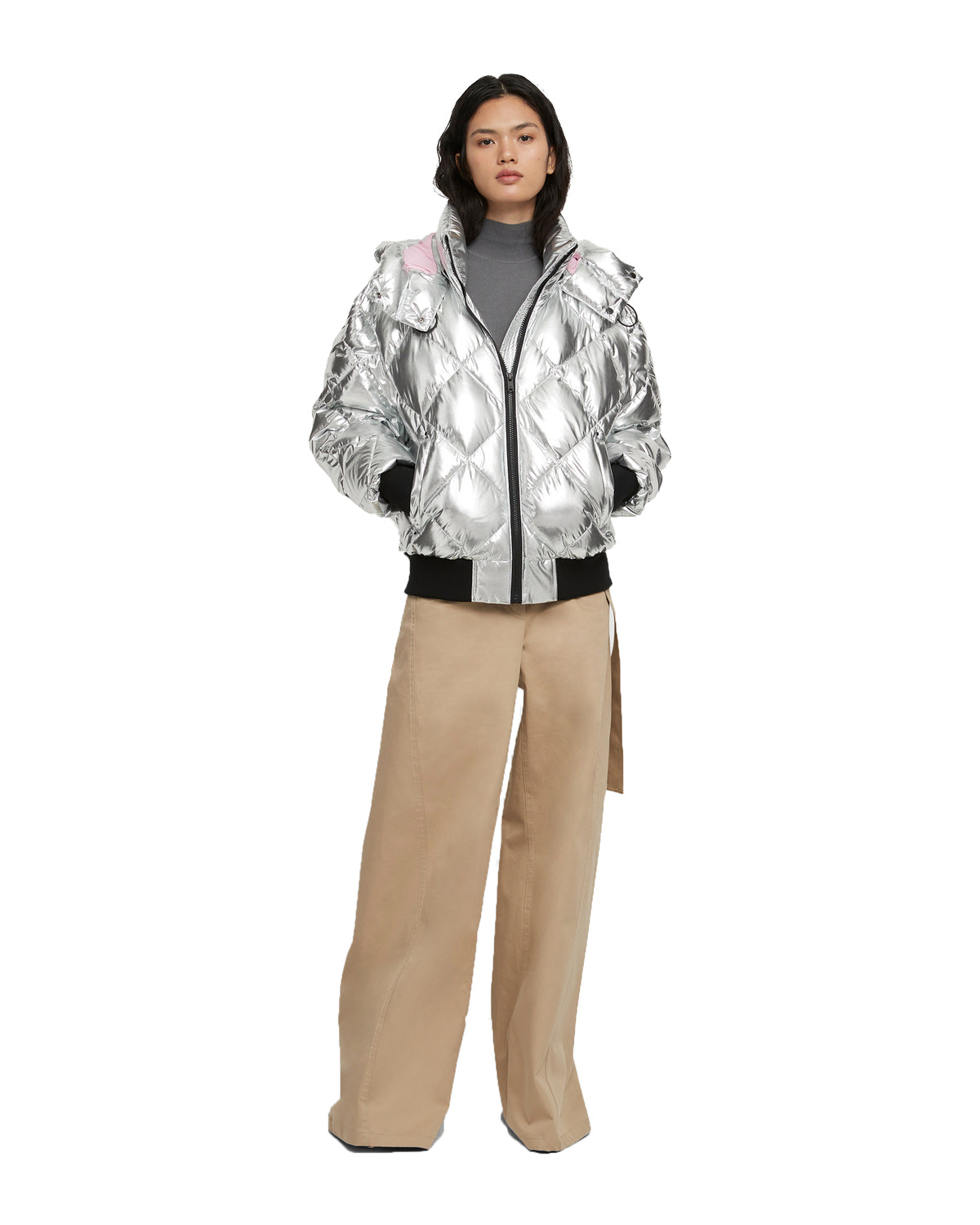 A woman standing wearing a silver quilted puffer jacket with a hood and wide-leg tan pants.