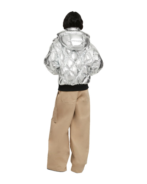 A person wearing a silver, quilted puffer jacket with a hood, paired with loose-fitting, light brown trousers.