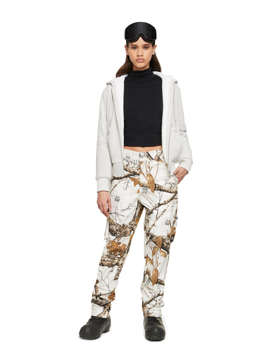 A person standing wearing goggles, black turtleneck, beige zip-up hoodie, and camouflage print pants.