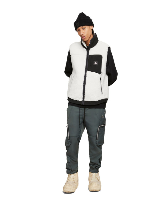 A person wearing a black beanie, black sweatshirt, white sherpa vest with a black pocket, dark green cargo pants, and tan boots, standing against a white background.