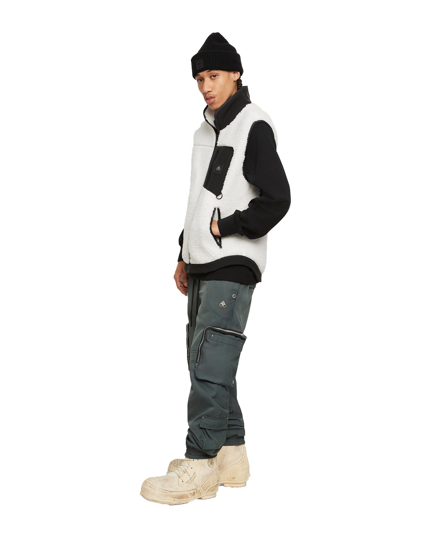 A person wearing a black beanie, black and white fleece vest, black turtleneck, dark green cargo pants, and light beige boots.