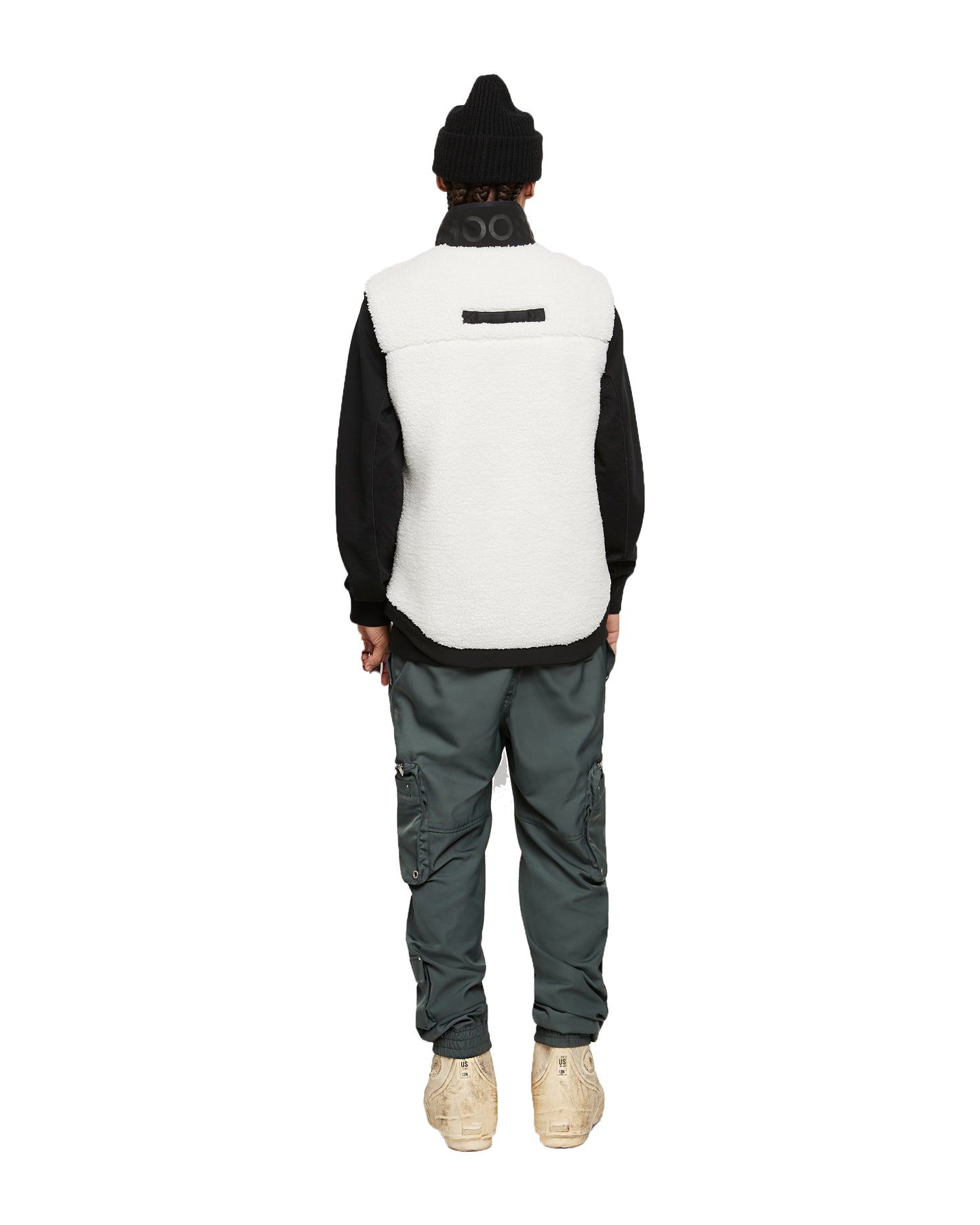 Back view of a person in a black knit cap, black long-sleeve shirt, a cream-colored fleece vest, and greenish-gray cargo pants and tan shoes.