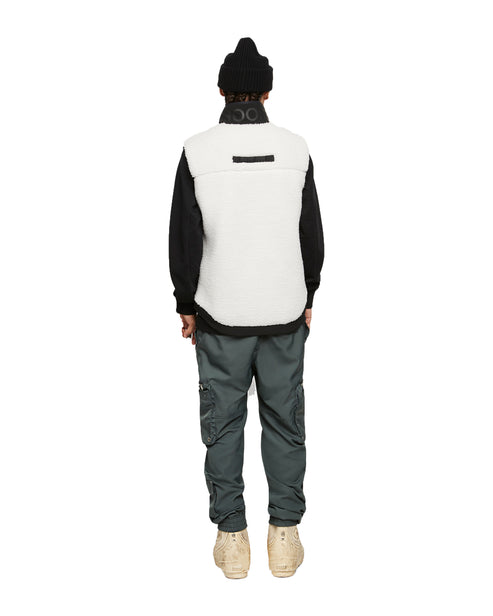 Back view of a person in a black knit cap, black long-sleeve shirt, a cream-colored fleece vest, and greenish-gray cargo pants and tan shoes.