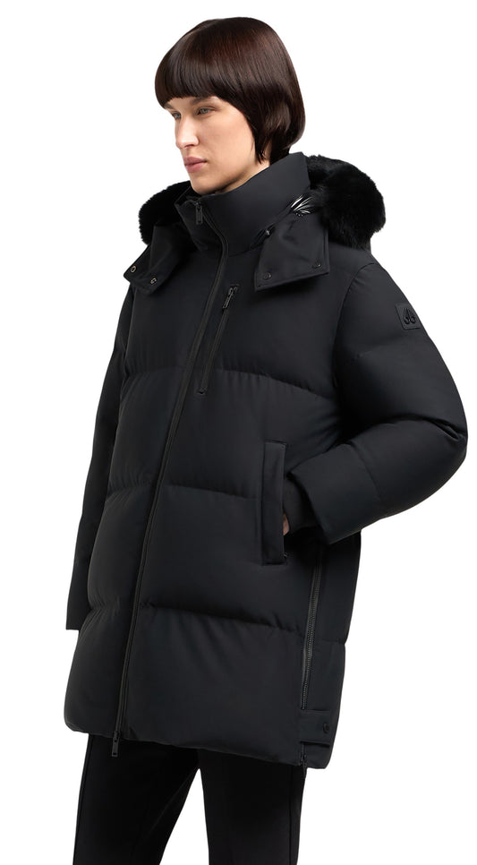 A person wearing a long, black puffer jacket with a fur-trimmed hood, the jacket features a zip closure and pockets.
