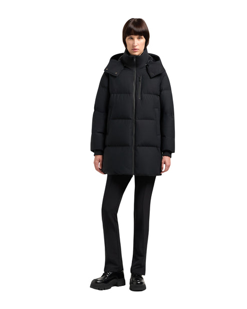 A woman is standing, wearing a long, black, hooded puffer coat and black pants with chunky shoes.