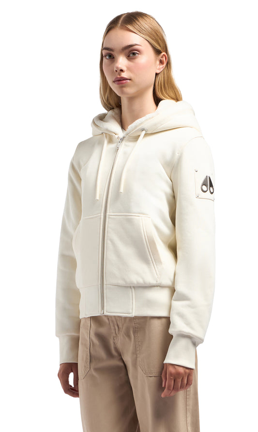 Woman wearing an off-white, zip-up hooded sweatshirt with a logo patch on the sleeve and matching colored pants.