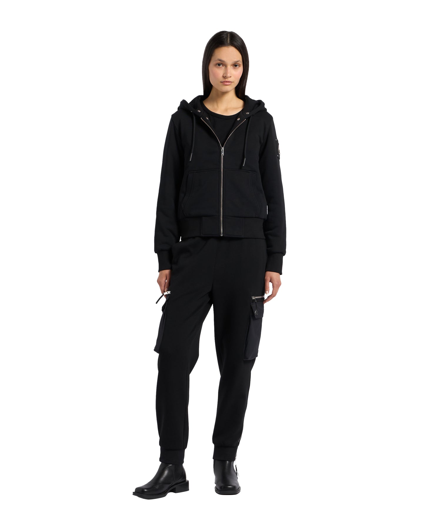 A person wearing a black hooded zip-up jacket and matching cargo pants stands against a white background.