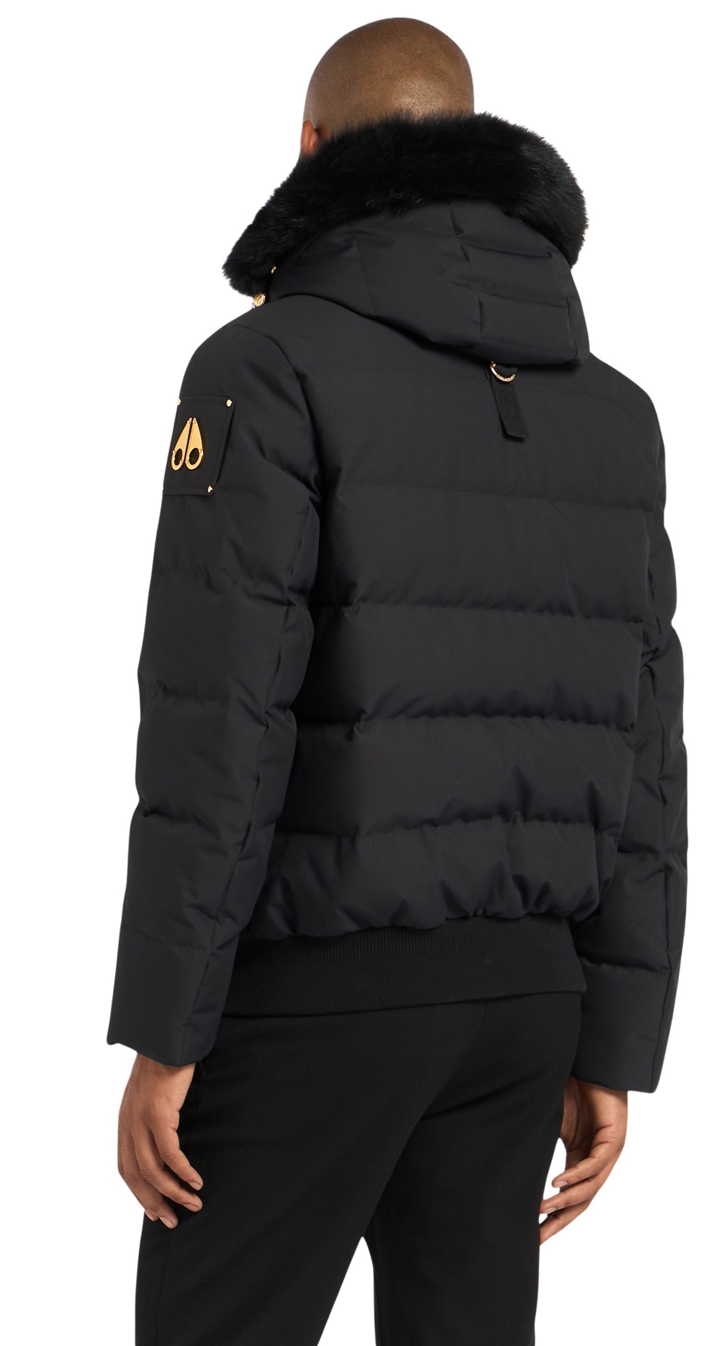 Back view of a black puffer jacket with a fur-trimmed hood, gold logo on the left sleeve, and ribbed hem.