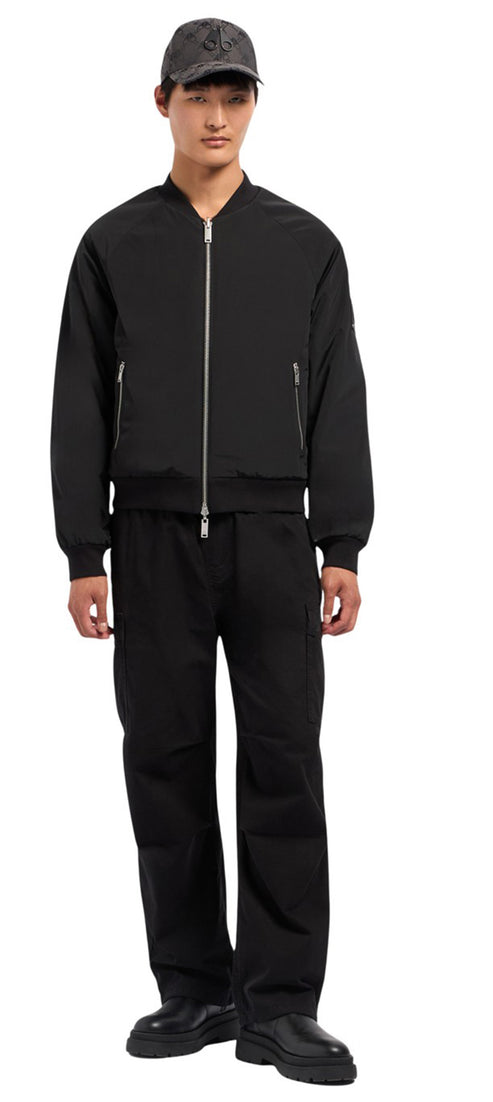 A person stands wearing a cap, a black bomber jacket, black pants, and black shoes.