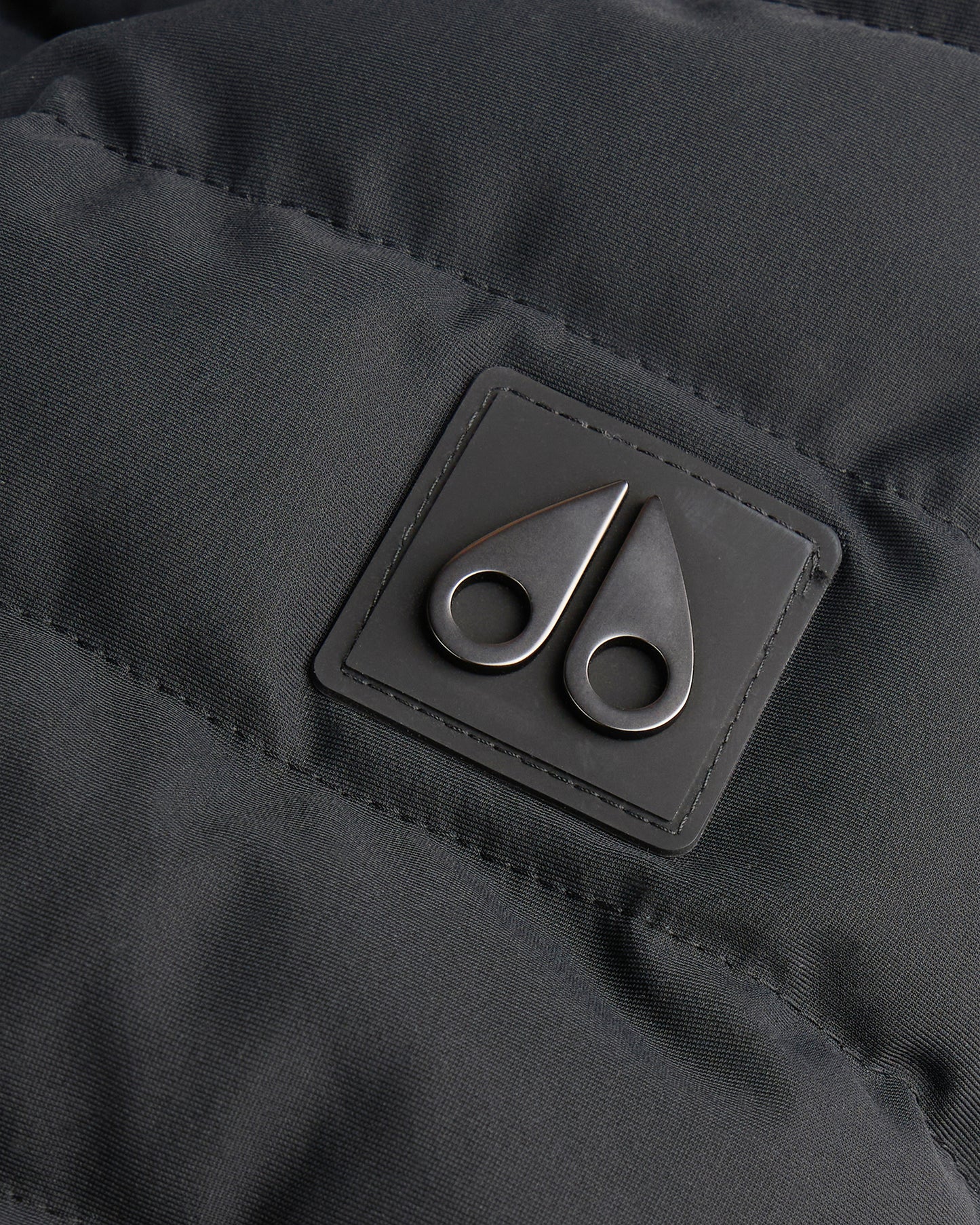 Close-up of the silver-colored logo consisting of two metal cleaver shapes on a dark gray stitched diamond-quilted material.