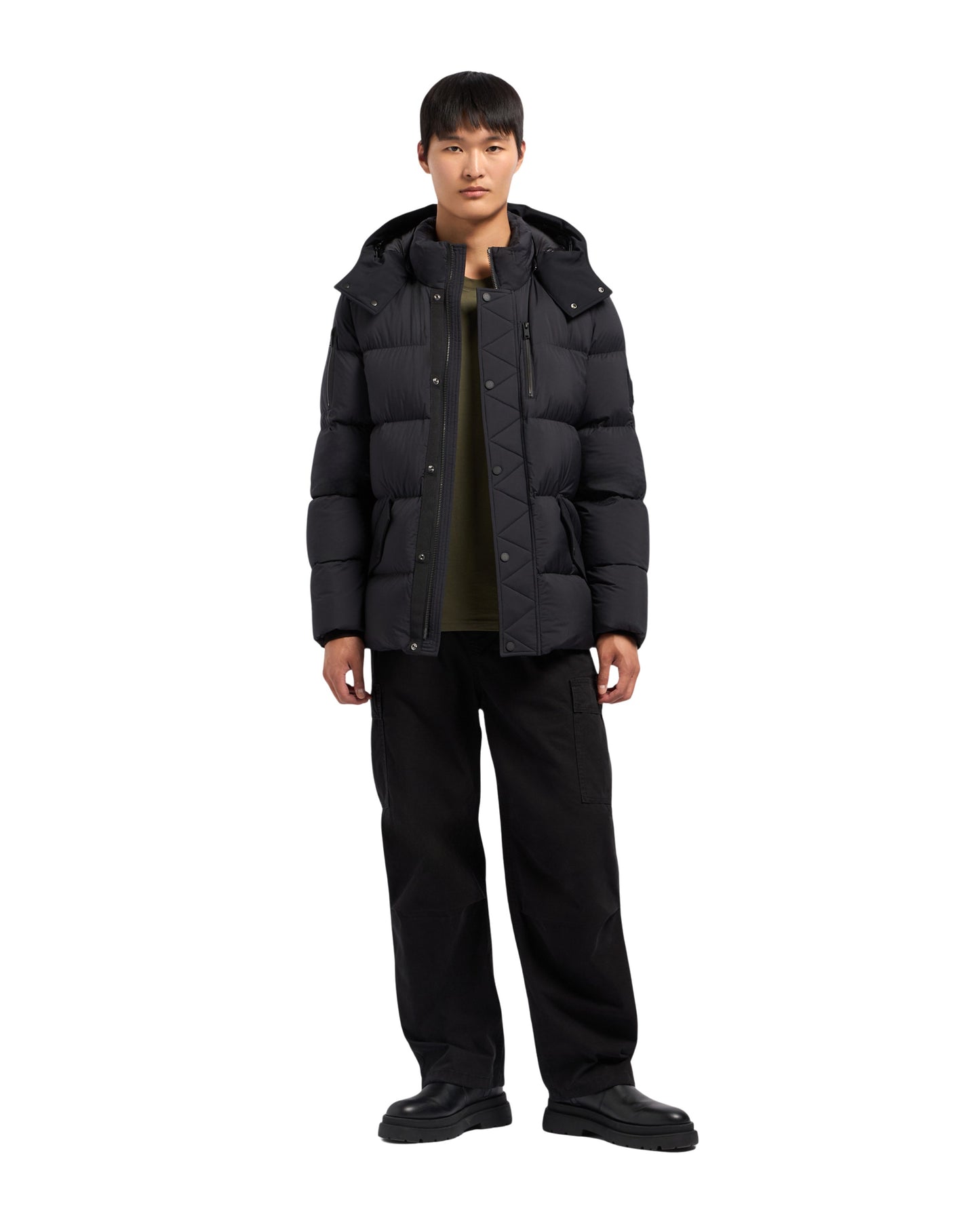 A person stands wearing a black puffer jacket with a hood, black pants, and black shoes.