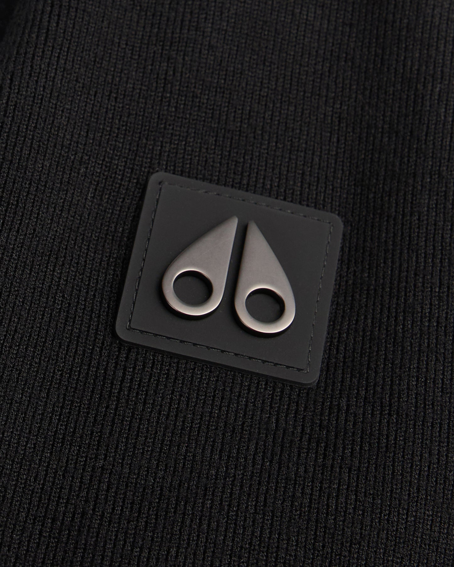 Close up of a black square leather patch with rounded edges, featuring two metallic, teardrop-shaped clasps or toggles pointing upward, stitched onto a textured black knit fabric.