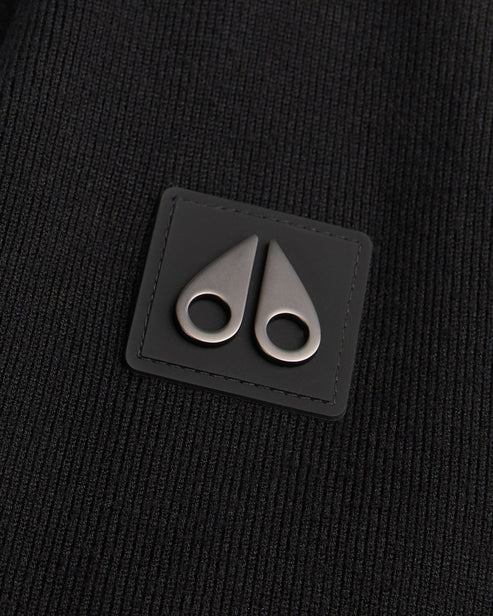 Close up of a black square leather patch with rounded edges, featuring two metallic, teardrop-shaped clasps or toggles pointing upward, stitched onto a textured black knit fabric.