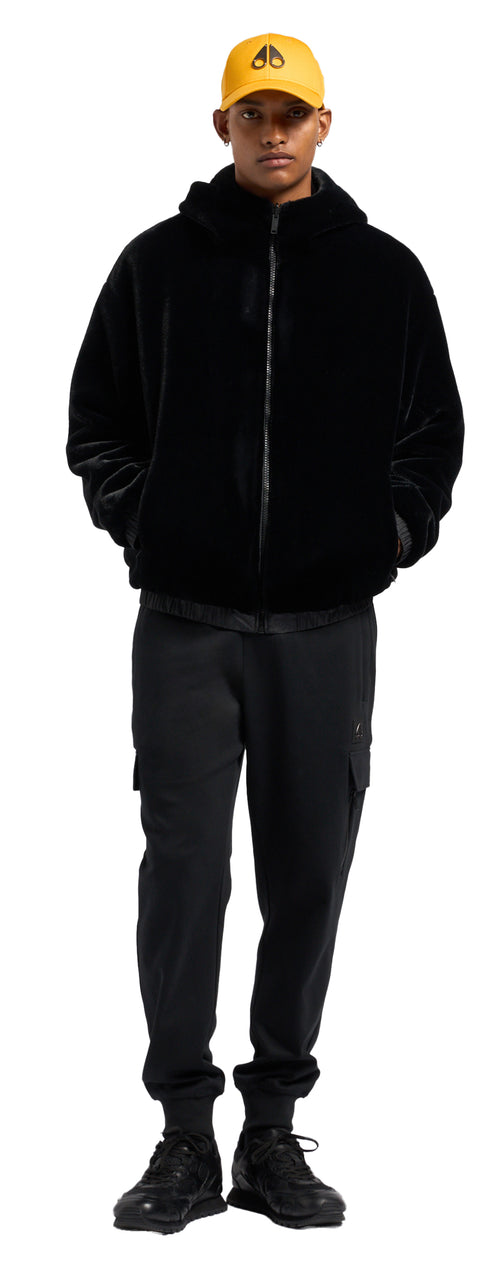 A man wearing a yellow cap with moose antlers logo, black jacket, black pants, and black shoes.