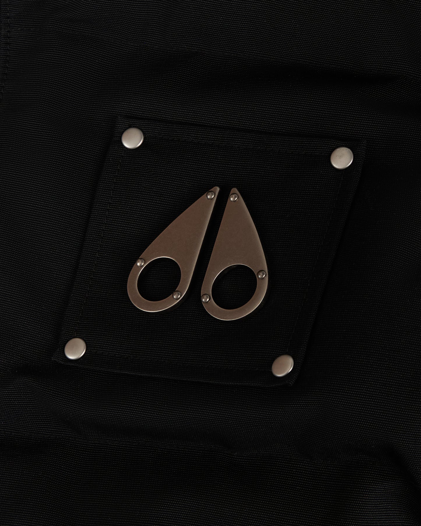 Black patch with metal logo resembling two teardrops, secured with four silver rivets.