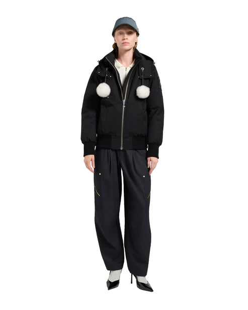 A woman in a black hooded jacket with two white pom poms, black pants and heels, wearing a blue hat.