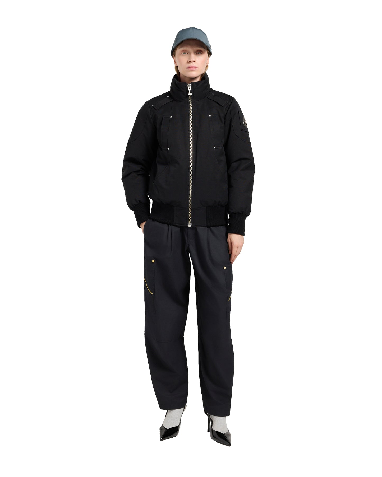 A person wears a black jacket, pants, heels, and a cap while standing.