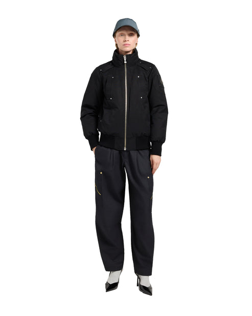 A person wears a black jacket, pants, heels, and a cap while standing.