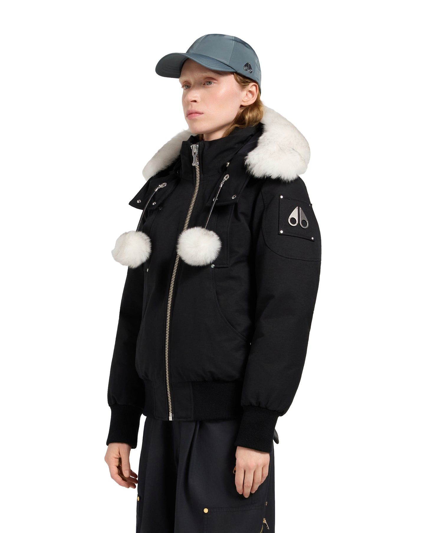 A woman wearing a black bomber jacket with white fur trim and two white pom poms attached to the chest, wearing a teal cap.