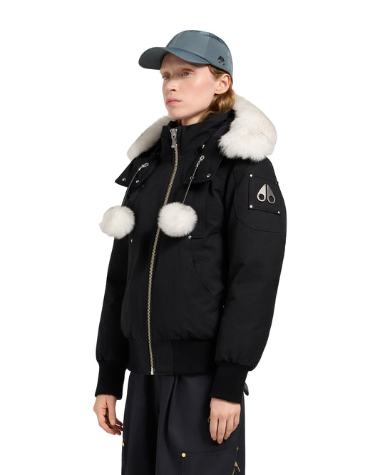 A woman wearing a black bomber jacket with white fur trim and two white pom poms attached to the chest, wearing a teal cap.