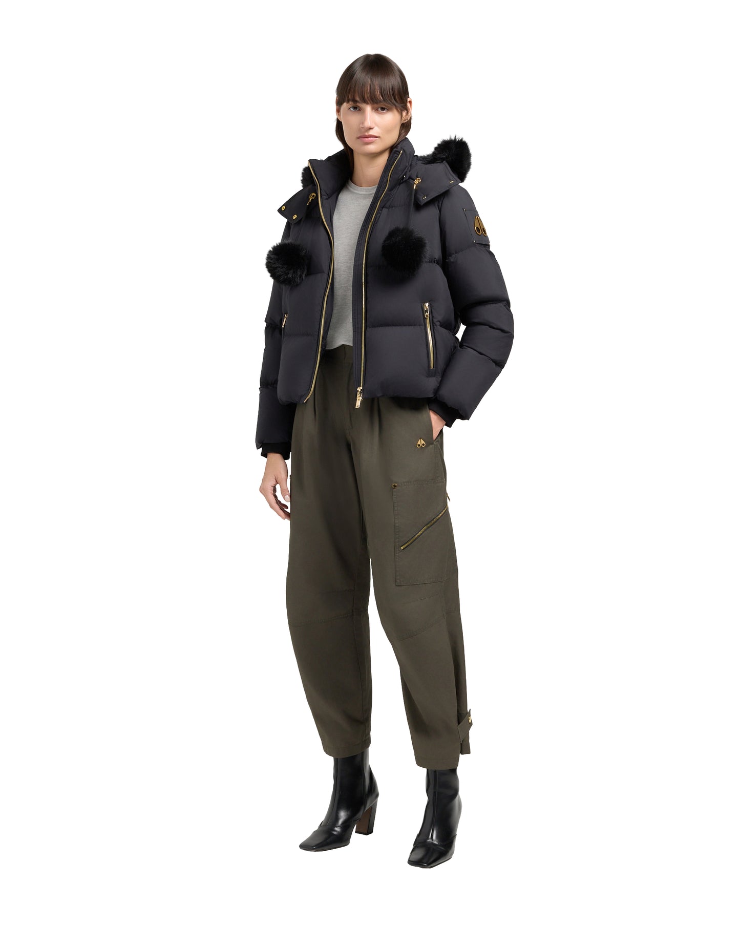 A person wearing a black puffer jacket with black fur pom poms on the shoulders and hood, a gray shirt, and olive green pants.