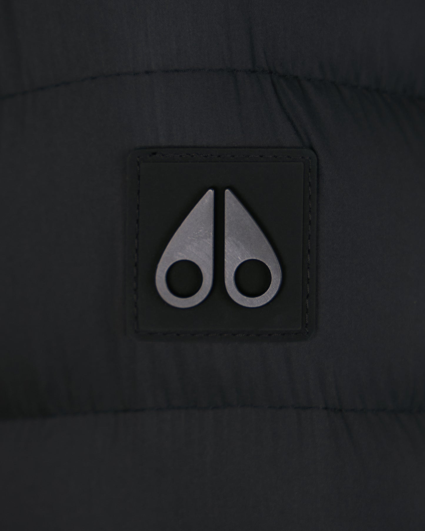 Close-up of a gray logo with two curved metal shapes resembling moose knuckles, stitched onto dark fabric.
