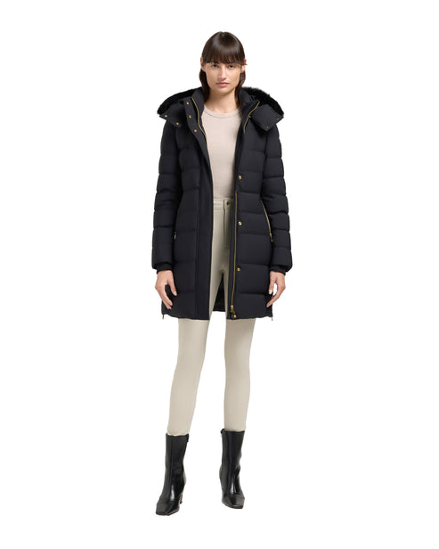 A woman wearing a black, mid-length puffer coat with a hood, light-colored pants, and black ankle boots.