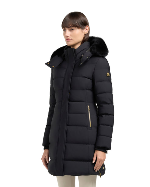 A woman poses in a black, knee-length puffer coat with a fur-trimmed hood and gold zippers.