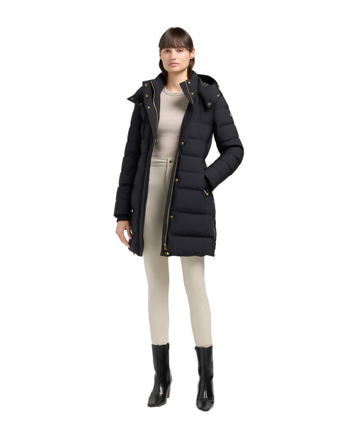 A woman stands wearing a black, long, hooded, puffer jacket with gold hardware.