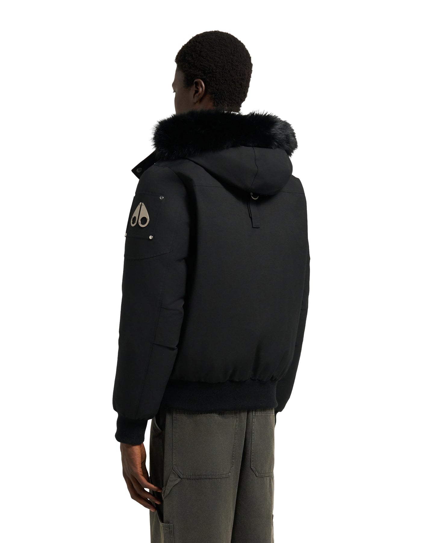 A man is seen from the back wearing a black bomber jacket with a fur-lined hood, and dark green pants.