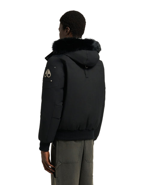 A man is seen from the back wearing a black bomber jacket with a fur-lined hood, and dark green pants.