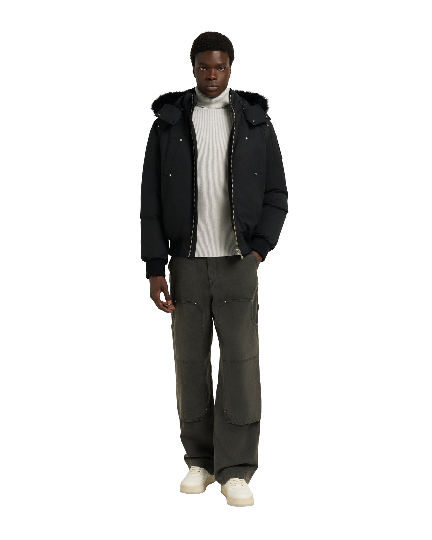 A dark-skinned male model wearing a black hooded down jacket, light grey turtleneck, wide leg dark green pants, and cream-colored shoes.