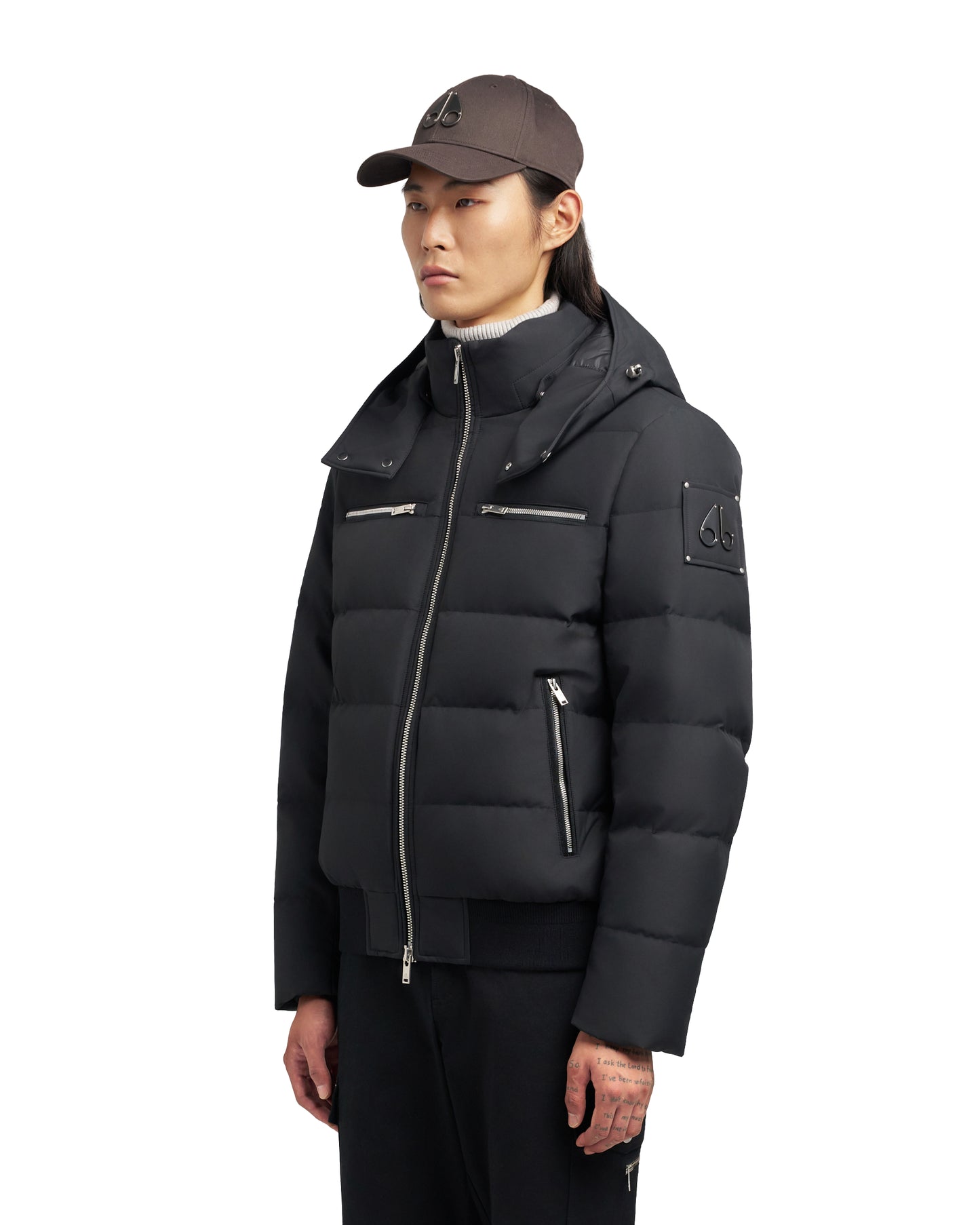 A person wears a dark bomber jacket with silver zippers and hood, plus a brown cap with a small logo.