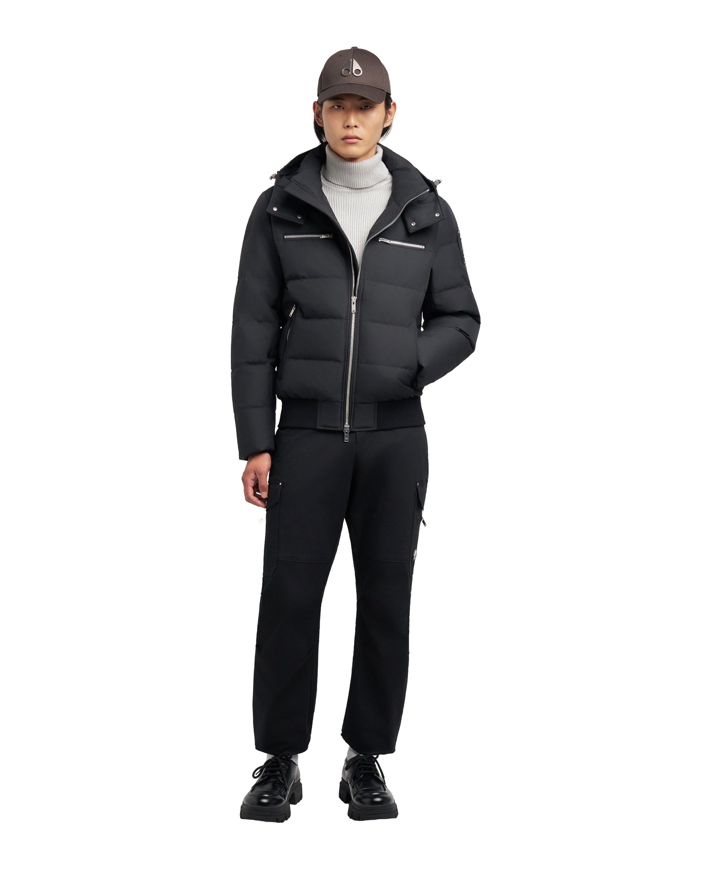 A person wearing a black puffer jacket with a hood, a grey turtleneck sweater, black pants, black shoes, and a brown cap.