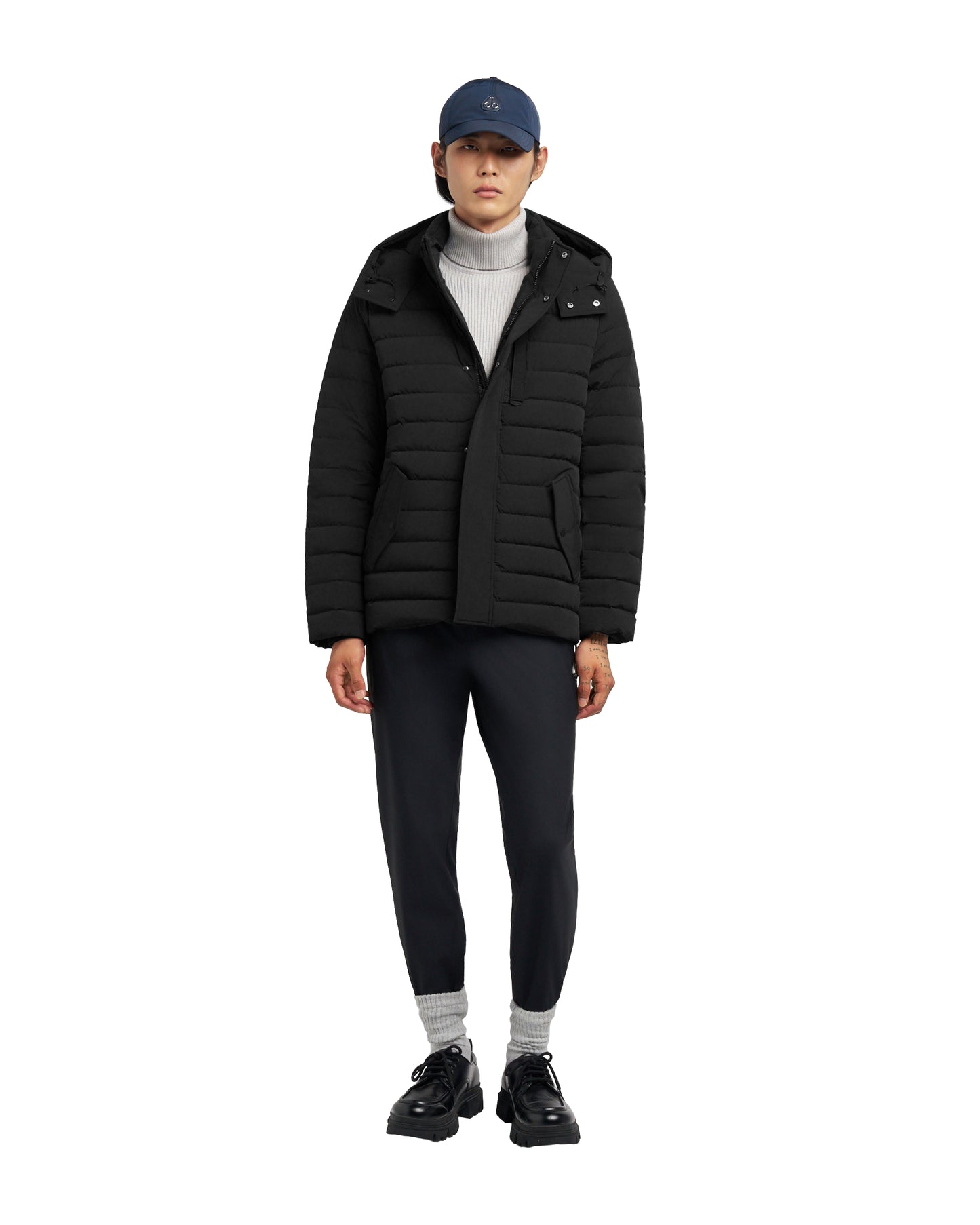 A person wearing a black puffer jacket, gray turtleneck, black pants and a blue cap.