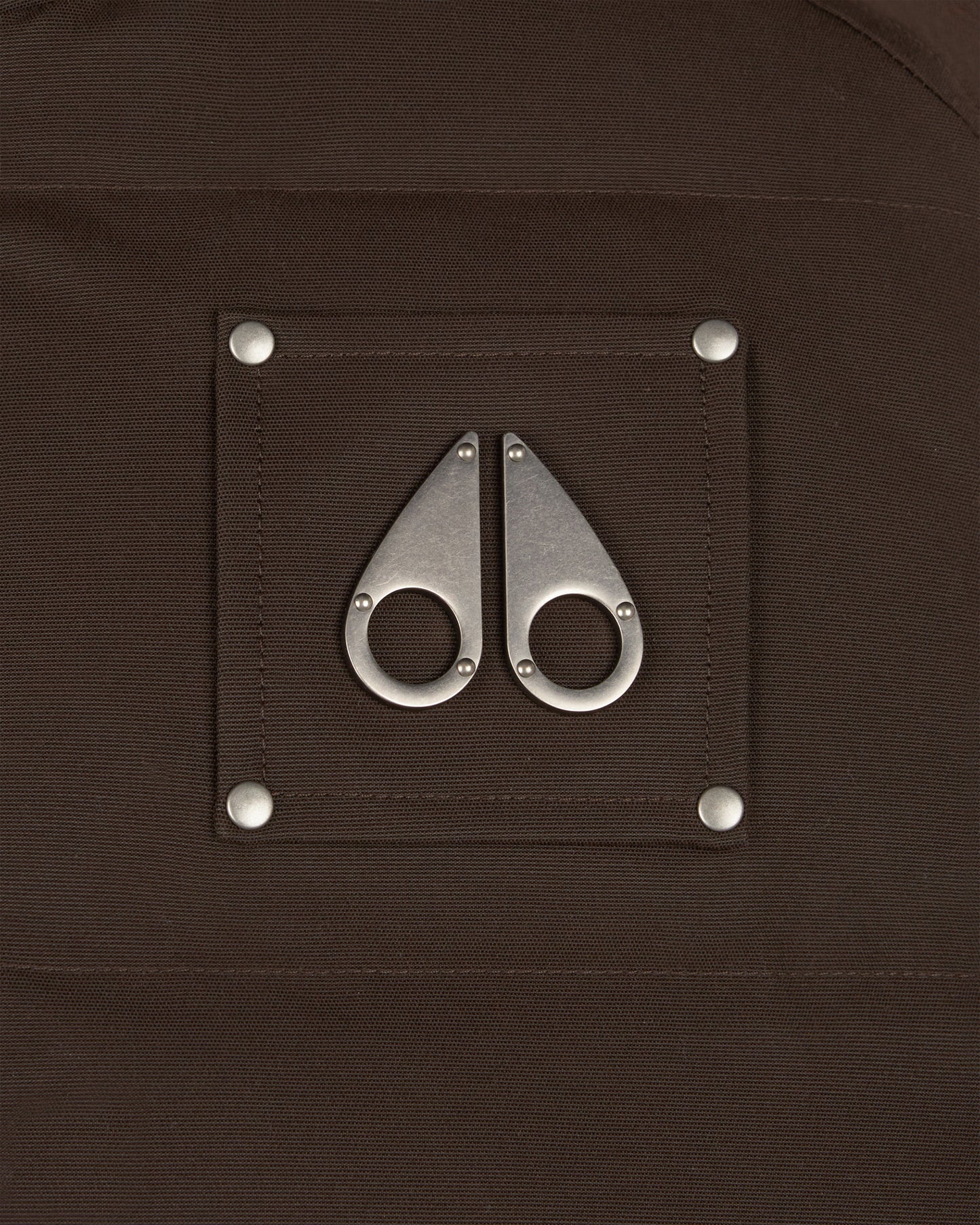 Close up of a brown fabric with metal logo, a square patch with two metal pieces that look like moose knuckles attached with silver rivets.