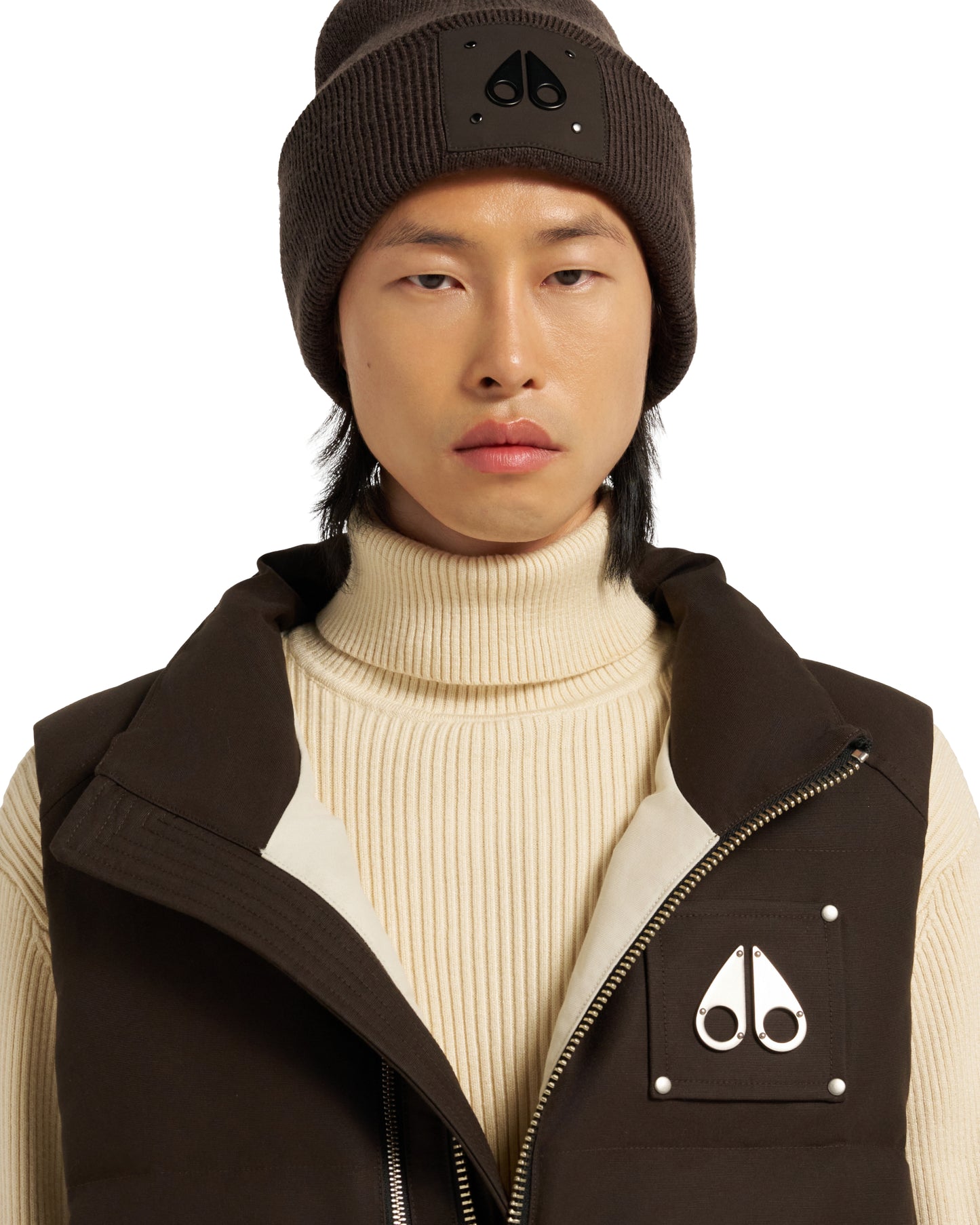 Person wearing brown beanie with logo, brown vest, and turtleneck sweater.