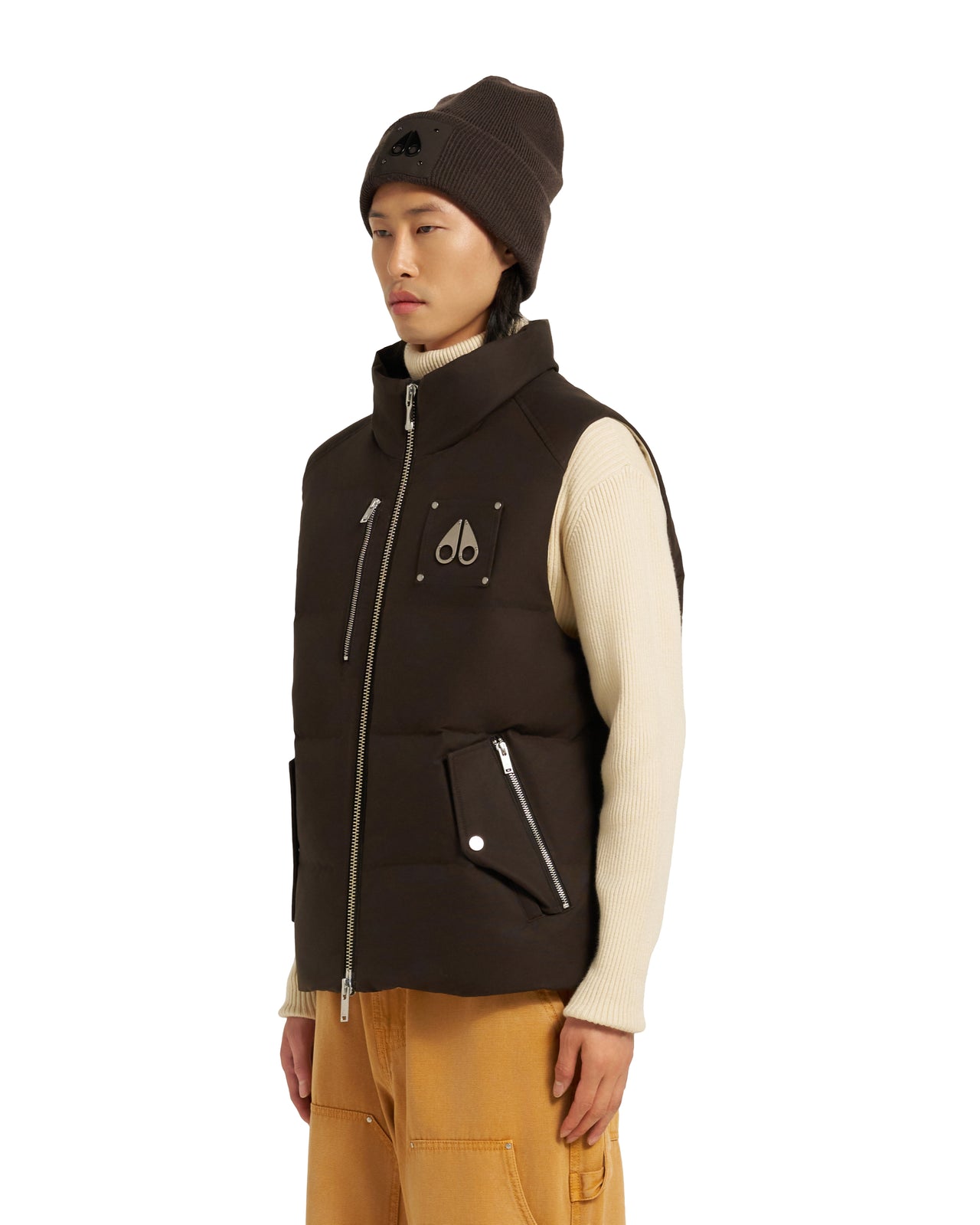 A person wearing a brown beanie, an off-white turtleneck sweater, a brown vest with a zipper on the front, and yellow pants.