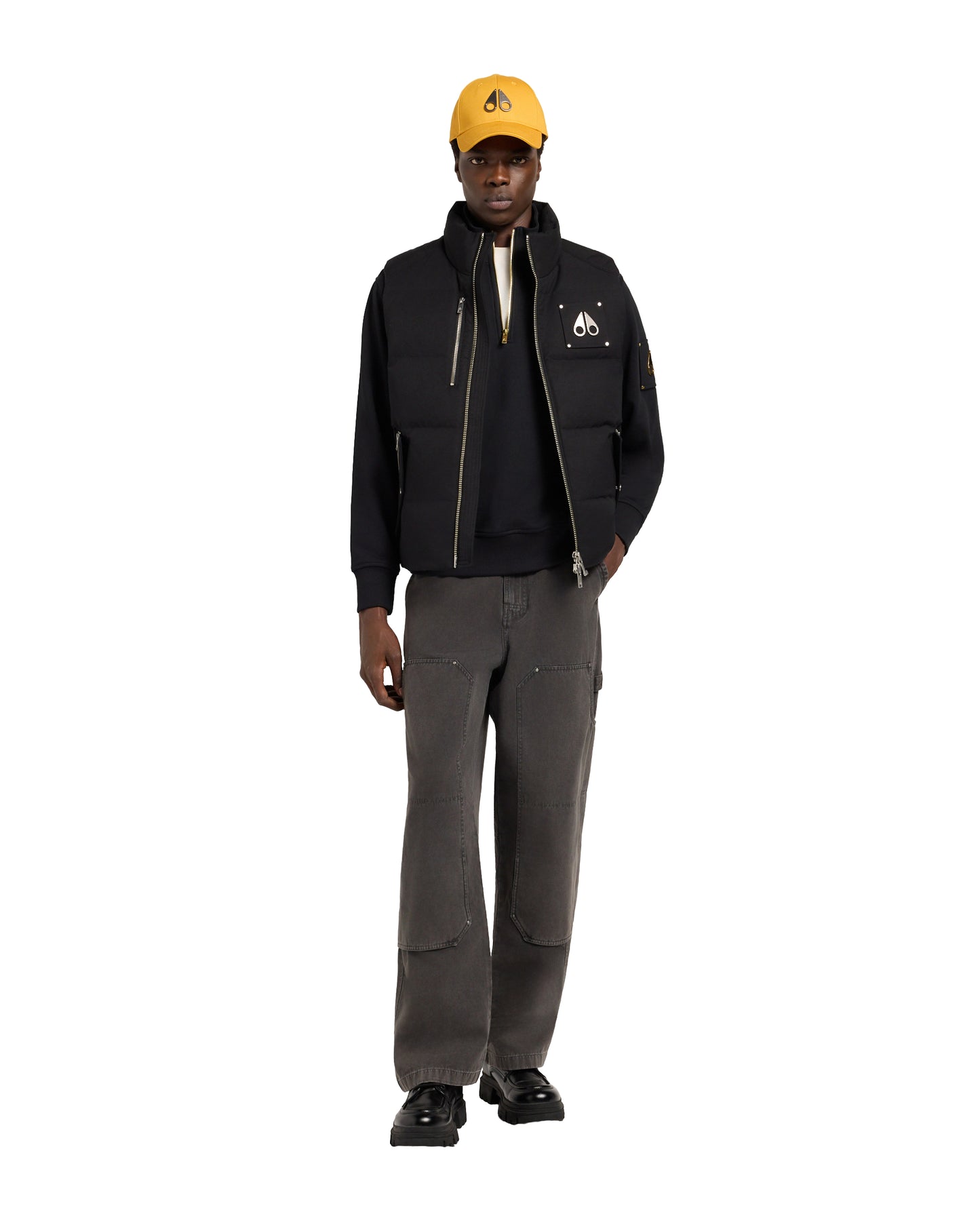 A man wearing a black zip-up vest with a high collar, paired with grey pants and a yellow baseball cap.