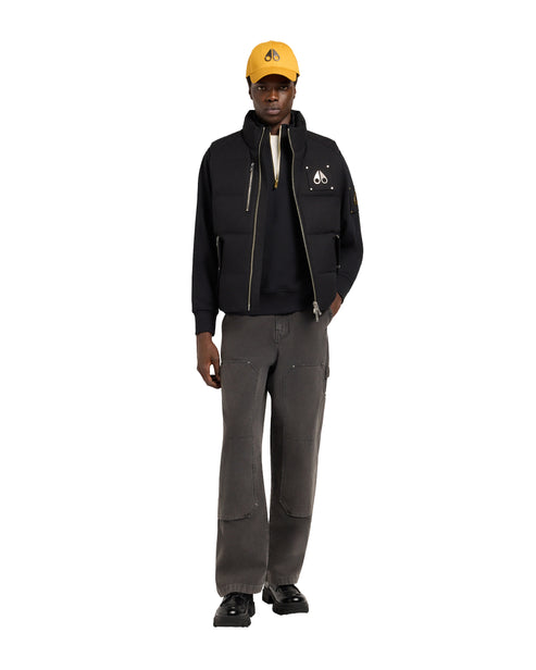 A man wearing a black zip-up vest with a high collar, paired with grey pants and a yellow baseball cap.