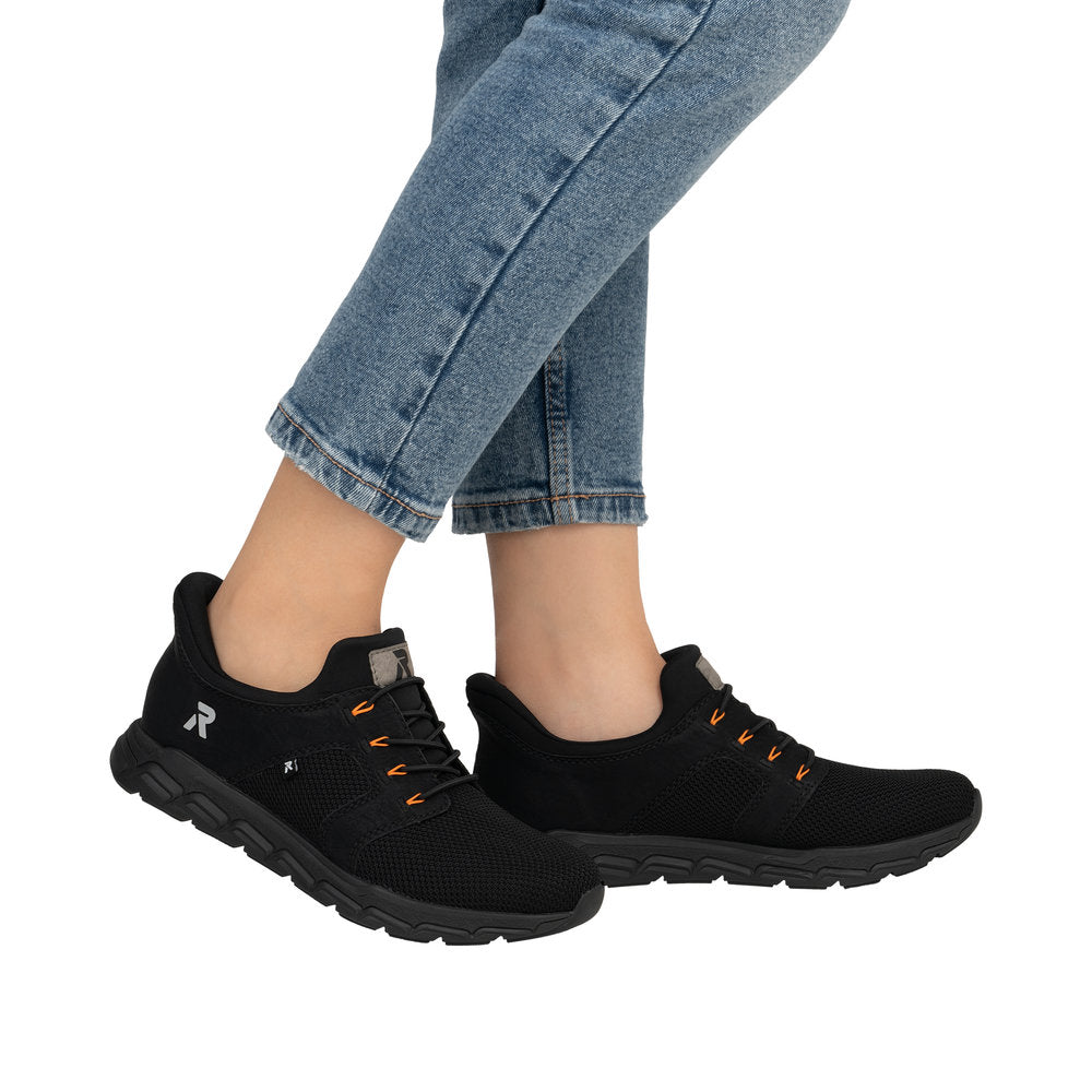 A pair of black athletic shoes with black laces that have orange accents, worn with blue jeans.