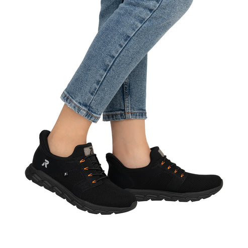 A pair of black athletic shoes with black laces that have orange accents, worn with blue jeans.