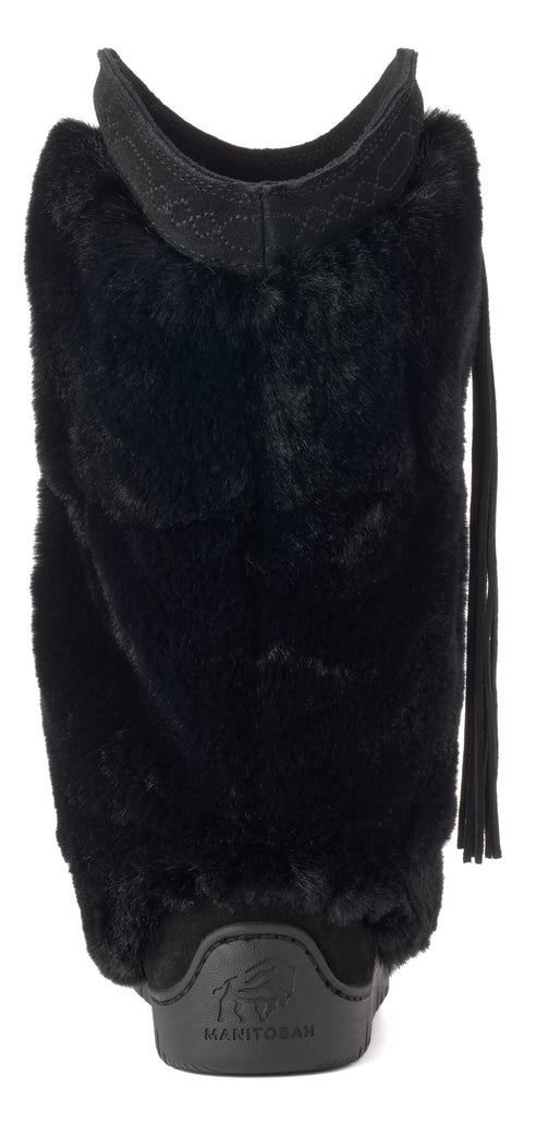 A black, mid-calf winter boot made of faux fur, featuring a black rubber heel base embossed with text, a wide black fabric strap at the top, and black fringe trim hanging from the right side.