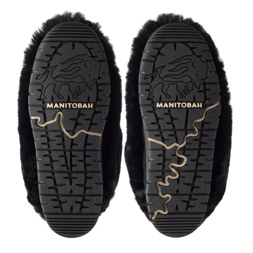 The black rubber outsoles of two fur-lined slippers, viewed from directly underneath. Each sole features a rugged tread pattern with a central stamped graphic of a buffalo silhouette and the word MANITOBAH, and a distinct, winding, gold-colored line inlay running along the side.