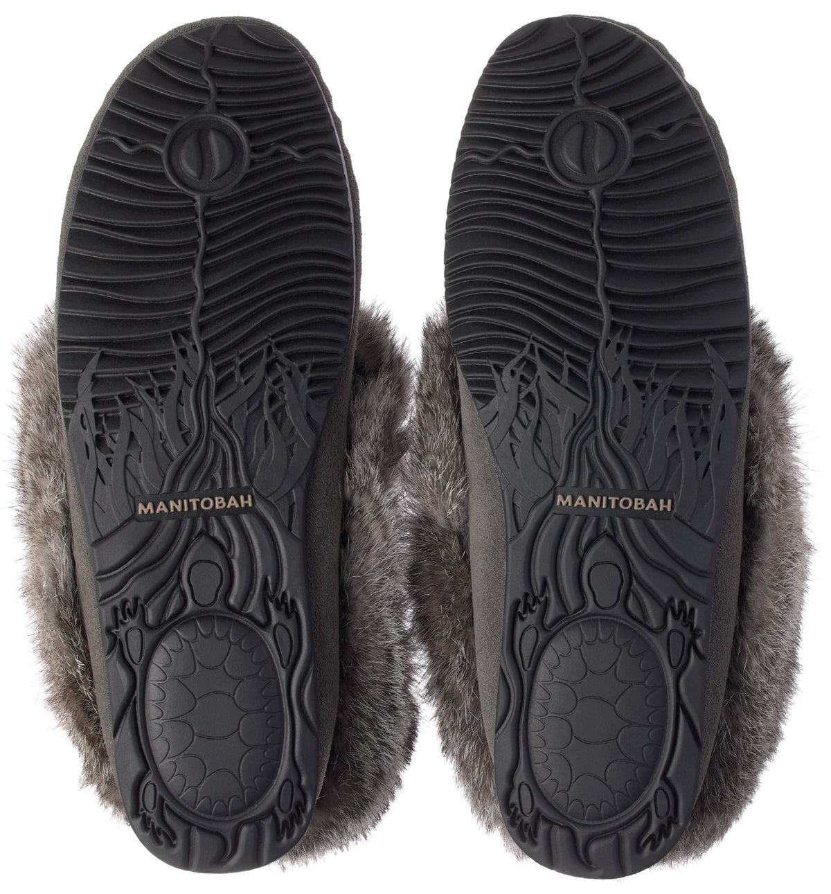 A pair of grey slippers viewed from the bottom, showing black rubber soles with a textured wave pattern, a turtle design at the heel, and the word MANITOBAH embossed in the center of the arch, surrounded by grey faux fur trim.