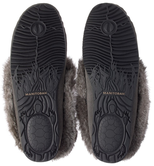 A pair of grey slippers viewed from the bottom, showing black rubber soles with a textured wave pattern, a turtle design at the heel, and the word MANITOBAH embossed in the center of the arch, surrounded by grey faux fur trim.