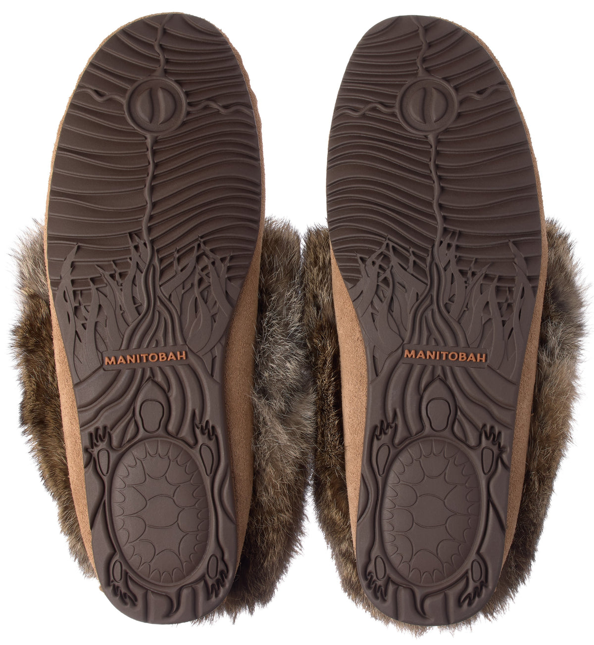 A pair of tan suede slippers with dark brown faux fur trim, shown from the bottom to display the molded rubber soles featuring a turtle graphic, a wave-like pattern, and the embossed name MANITOBAH.